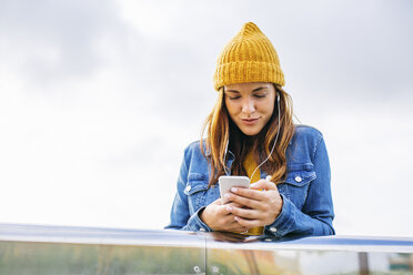 Lächelnde junge Frau mit gelber Mütze, die auf ihr Mobiltelefon schaut - EBSF001684