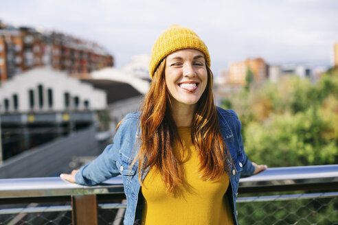 Porträt einer lächelnden jungen Frau mit gelber Mütze, die ihre Zunge herausstreckt - EBSF001692