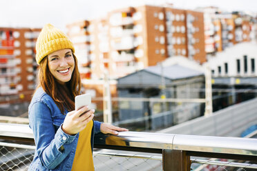 Porträt einer lächelnden jungen Frau mit Mobiltelefon und gelber Kappe - EBSF001694
