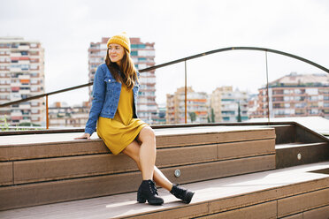 Smiling young woman wearing yellow cap and dress sitting on steps - EBSF001695