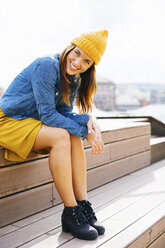 Porträt einer lächelnden jungen Frau mit gelber Mütze, die auf einer Treppe sitzt - EBSF001696