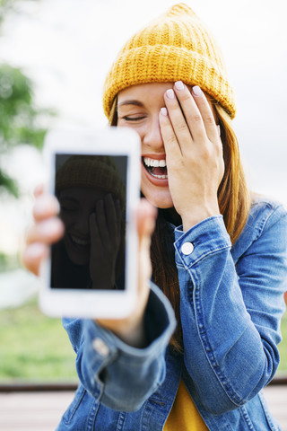 Porträt einer lachenden jungen Frau, die ein Smartphone mit ihrem eigenen Bild zeigt, lizenzfreies Stockfoto