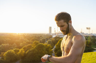 Barechested athlete with earbuds looking on watch at sunset - DIGF001115