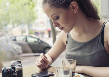Junge Frau in einem Café mit Kamera, die ihr Mobiltelefon überprüft - TAMF000602