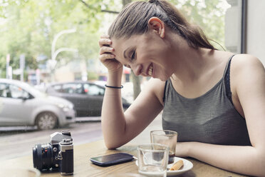 Lächelnde junge Frau in einem Café mit Kamera, die ihr Mobiltelefon überprüft - TAMF000603