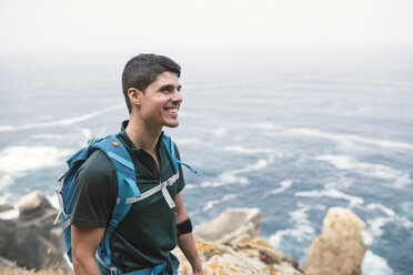 Lächelnder Mann mit einem Rucksack vor dem Meer an der Küste - RAEF001448