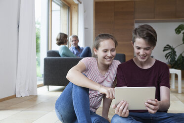 Lächelnde Teenager-Geschwister teilen sich ein Tablet im Wohnzimmer mit den Eltern im Hintergrund - RBF005166