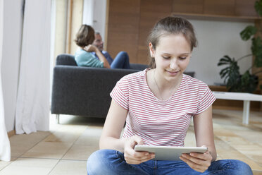 Lächelndes Mädchen mit Tablet im Wohnzimmer mit Eltern im Hintergrund - RBF005168