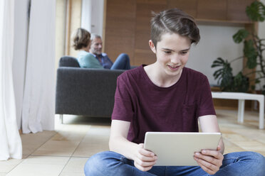 Smiling teenage boy using tablet in living room with parents in background - RBF005169