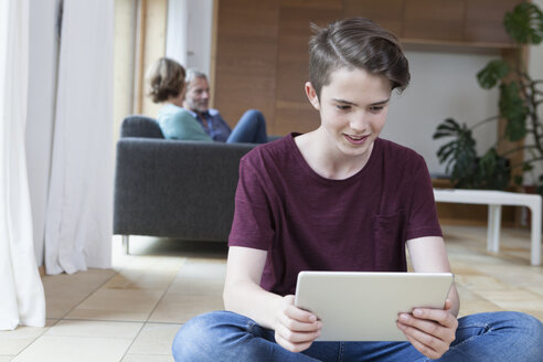 Lächelnder Teenager, der ein Tablet im Wohnzimmer benutzt, mit seinen Eltern im Hintergrund - RBF005169