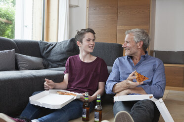 Vater und Sohn sitzen auf dem Boden und essen Pizza im Wohnzimmer - RBF005173