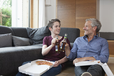 Father and son sitting on the floor clinking bottles in living room - RBF005174