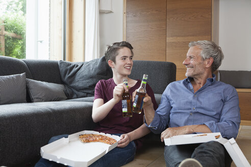 Vater und Sohn sitzen auf dem Boden und stoßen im Wohnzimmer mit Flaschen an - RBF005174