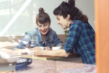 Two women sitting behind windowpane of a coffee shop looking at smartphone - TSFF000082