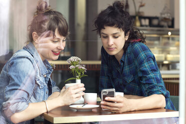 Zwei Frauen sitzen hinter der Fensterscheibe eines Cafés und schauen auf ihr Smartphone - TSFF000083