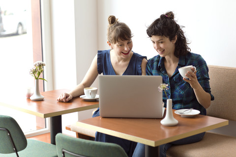 Zwei lächelnde Freunde sitzen in einem Café und schauen auf einen Laptop, lizenzfreies Stockfoto