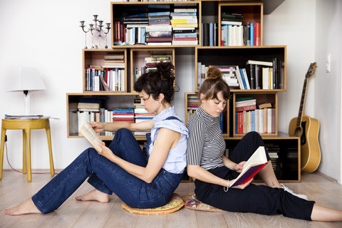 Zwei lesende Freunde sitzen Rücken an Rücken auf dem Boden des Wohnzimmers, lizenzfreies Stockfoto