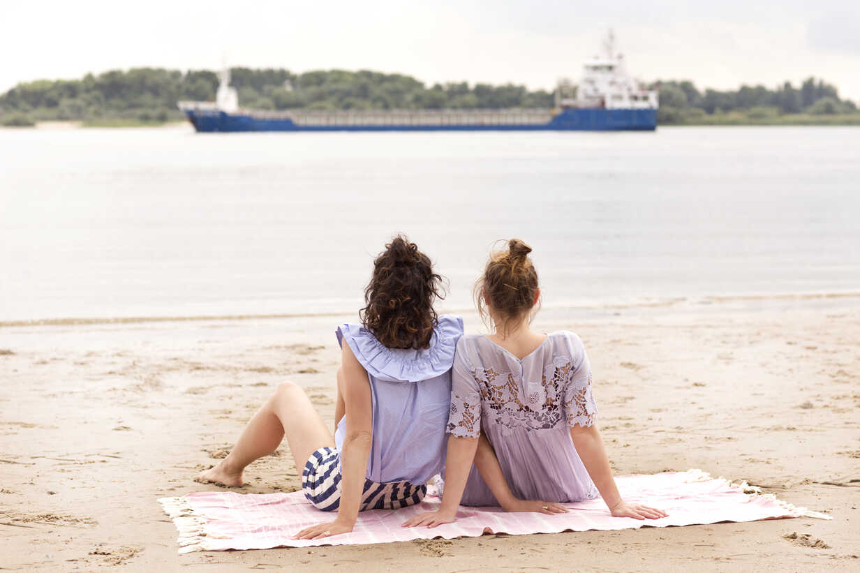 Back view of two friends sitting side by side on the beach watching a ship  stock photo