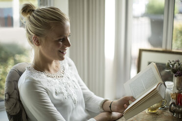 Lächelnde junge Frau, die am Fenster ein Buch liest - ZEF010062