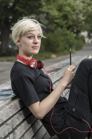 Porträt einer jungen Frau mit Kopfhörern, die auf einer Parkbank sitzt, lizenzfreies Stockfoto