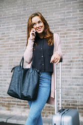 Smiling young woman with wheeled luggage and leather bag on the phone - EBSF001715