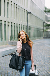 Smiling young woman with wheeled luggage and leather bag on the phone - EBSF001717