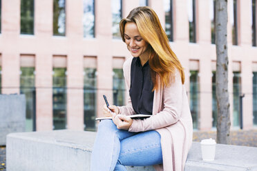 Lächelnde junge Frau auf einer Bank sitzend mit Notebook und Kaffee zum Mitnehmen - EBSF001719