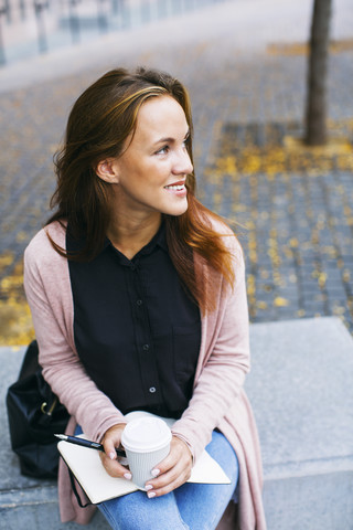 Lächelnde junge Frau auf einer Bank sitzend mit Notebook und Kaffee zum Mitnehmen, lizenzfreies Stockfoto