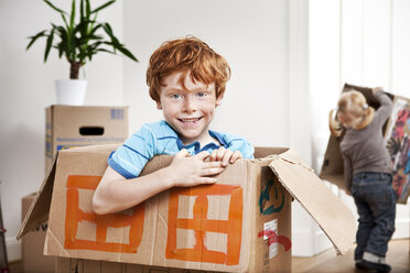 Glücklicher Junge schaut aus einem Karton in seiner neuen Wohnung mit seiner Schwester im Hintergrund - MFF003123