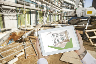 Hands holding a digital tablet with an architectural house sketch in front of a contruction site - MFF003298