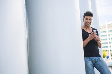 Smiling young man with smartphone leaning against column - SIPF000836