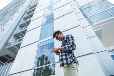 Junger Mann steht vor einem Hochhaus und schaut auf sein Handy - SIPF000838