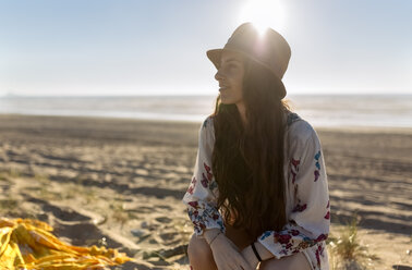 Lächelnde junge Frau sitzt am Strand im Gegenlicht - MGOF002426