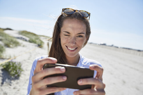 Lächelnde junge Frau blinzelt mit Handy in der Hand am Strand - SRYF000070