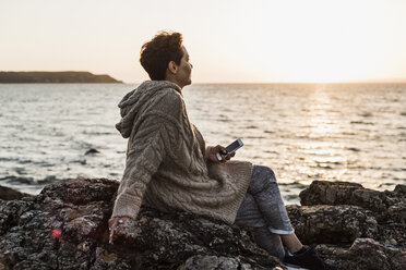 Frankreich, Halbinsel Crozon, Frau mit Smartphone auf Felsen sitzend bei Sonnenuntergang - UUF008340