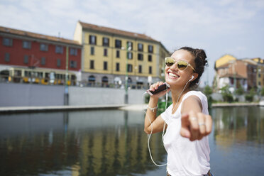 Happy young woman listening to music at the riverside - MRAF000173