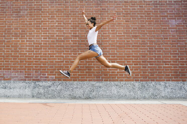 Übermütige junge Frau, die vor einer Backsteinmauer springt - MRAF000176
