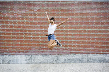 Exuberant young woman jumping in front of a brick wall - MRAF000177