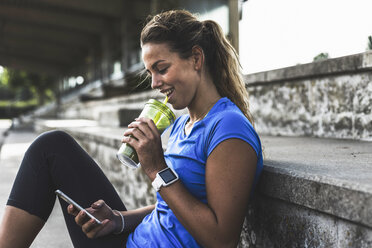 Sportive young woman sitting on grandstand with cell phone and drinking mug - UUF008384