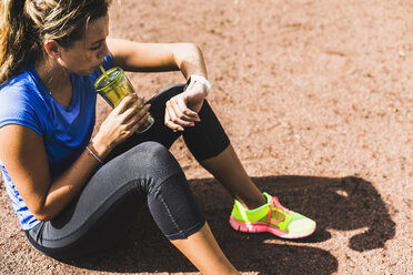 Sportliche junge Frau sitzt auf einem Sportplatz, trinkt aus einem Becher und schaut auf ihre Smartwatch - UUF008391