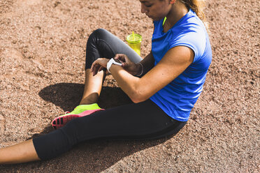 Sportliche junge Frau sitzt auf einem Sportplatz und schaut auf ihre Smartwatch - UUF008392