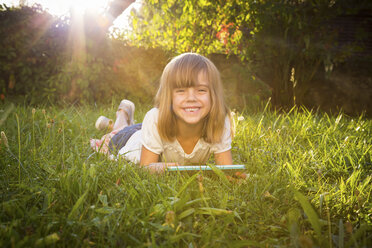 Porträt eines grinsenden kleinen Mädchens auf einer Wiese liegend mit Tablet - LVF005286