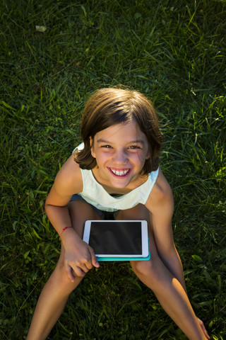 Porträt eines lächelnden Mädchens, das mit einem Tablet auf einer Wiese sitzt und zur Kamera aufschaut, lizenzfreies Stockfoto