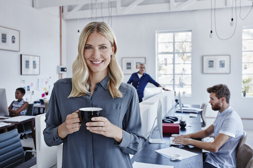 Porträt einer lächelnden Geschäftsfrau im Büro mit Mitarbeitern im Hintergrund - RORF00290