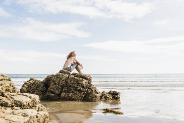 Teenager-Mädchen sitzt auf einem Felsen am Strand und hört Musik - UUF08404