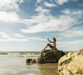 Teenage girl sitting on rock on the beach taking a selfie - UUF08406