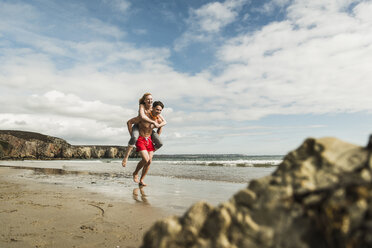 Happy young man carrying girlfriend piggyback on the beach - UUF08409