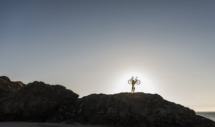 Frankreich, Halbinsel Crozon, Mountainbiker hebt sein Fahrrad bei Sonnenuntergang hoch - UUF08509