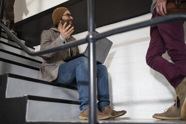 Hipper Kreativprofi mit Laptop auf einer Treppe sitzend, im Gespräch mit einem Kollegen - ZEF10376