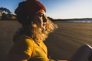 Teenage girl wearing beanie and headphones sitting on the beach - UUF08533